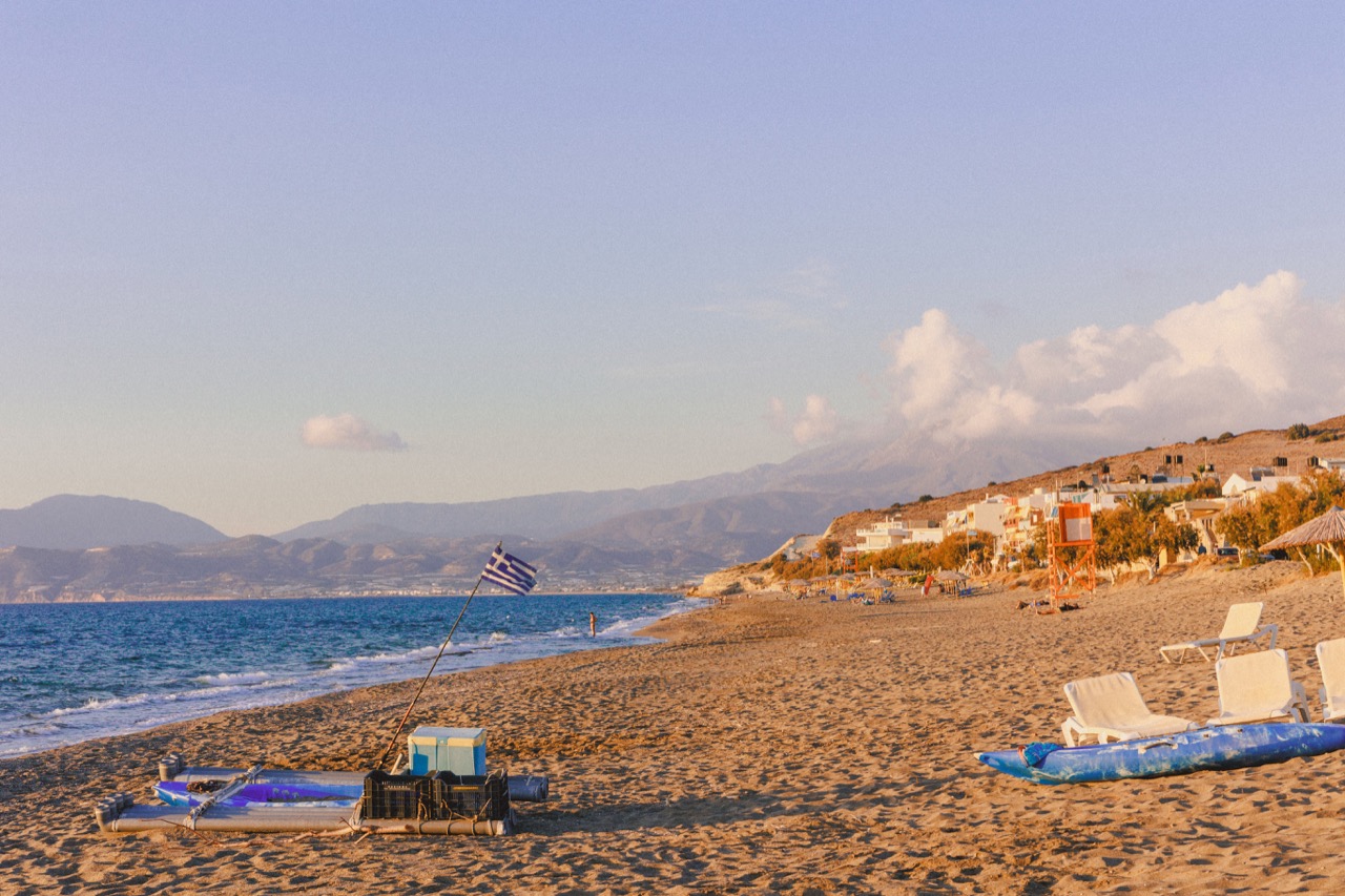 A sandy beach with sunbeds and a Greek flag, mountains across the water